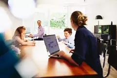 Mann und Frau arbeiten an Laptop und PC und sprechen lächelnd miteinander, während Kinder am Tisch zeichnen